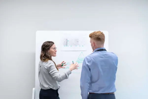 Two people stand in front of white board and discuss the pros and cons of hiring