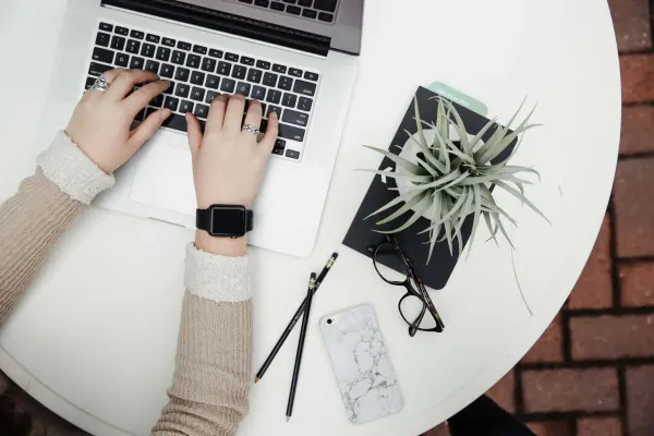 A blogger types at a laptop on a white tabletop, their smartphone next to them,