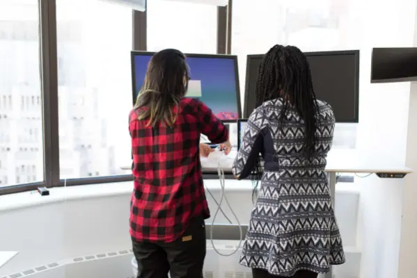Two digital marketers stand at a desk in front of multiple monitors, discussing
