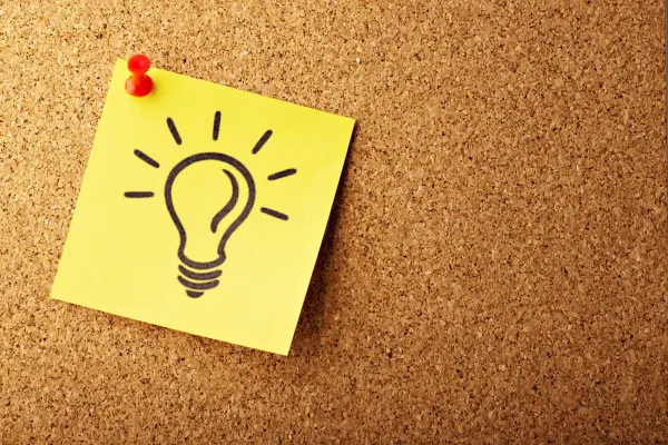 A yellow post it note with a light bulb is pinned to a cork board